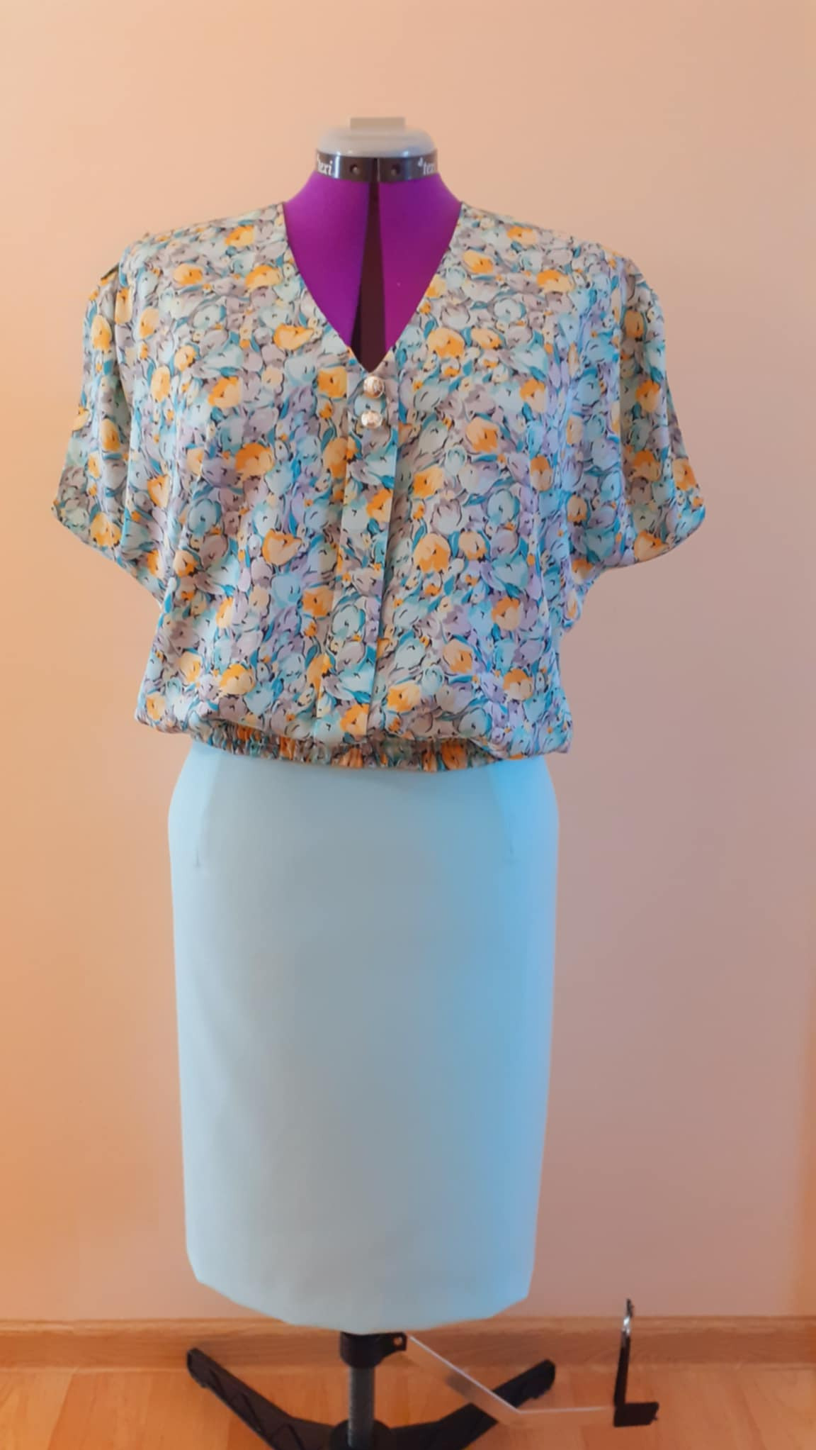 Damska bluzka w kwiaty z krótkim rękawem i jasnoniebieska spódnica ołówkowa na manekinie krawieckim z fioletową szyją.