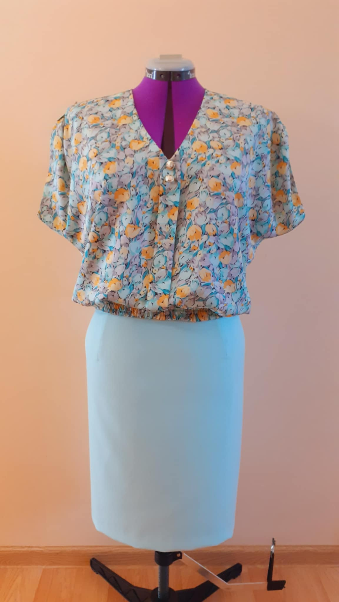 Bluzka z krótkim rękawem w kwiatowy wzór, zapinana na dwa guziki, zestawiona z dopasowaną spódnicą na manekinie krawieckim z fioletowym kołnierzem.
