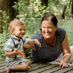 Uśmiechnięta kobieta w bluzce w paski i białe spodnie oraz mały chłopiec z szelkami siedzą na drewnianym podwyższeniu w lesie. Chłopiec trzyma kawałek arbuza.