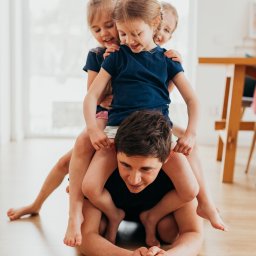 Weronika Zelewska Fotografia-Usługi Fotograficzne - Ojciec leżący na podłodze, na nim siedzą trzy uśmiechnięte dziewczynki w identycznych granatowych koszulkach, jasne wnętrze z drewnianą podłogą.