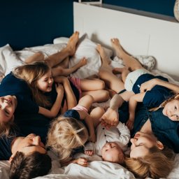Weronika Zelewska Fotografia-Usługi Fotograficzne - Duża rodzina leży na łóżku, rodzice i dzieci, uśmiechnięci, w niebieskich ubraniach, widok z góry, na białej pościeli.