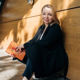Weronika Zelewska Fotografia-Usługi Fotograficzne - Uśmiechnięta blondynka w czarnym żakiecie, siedząca na drewnianej podłodze przy ścianie z okładziną, trzymająca pomarańczową książkę 'Teatr Umysłu' z czerwoną grafiką.