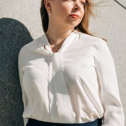 Weronika Zelewska Fotografia-Usługi Fotograficzne - Portret kobiety w białej bluzce i granatowej spódnicy, oparta o szarą ścianę w słoneczny dzień, wiatr rozwiewa jej włosy.