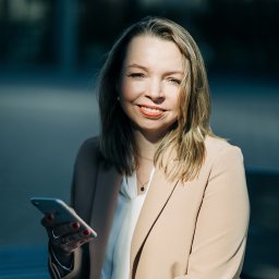 Weronika Zelewska Fotografia-Usługi Fotograficzne - Portret uśmiechniętej kobiety w beżowym żakiecie, trzymającej smartfon na tle miejskiego pejzażu.