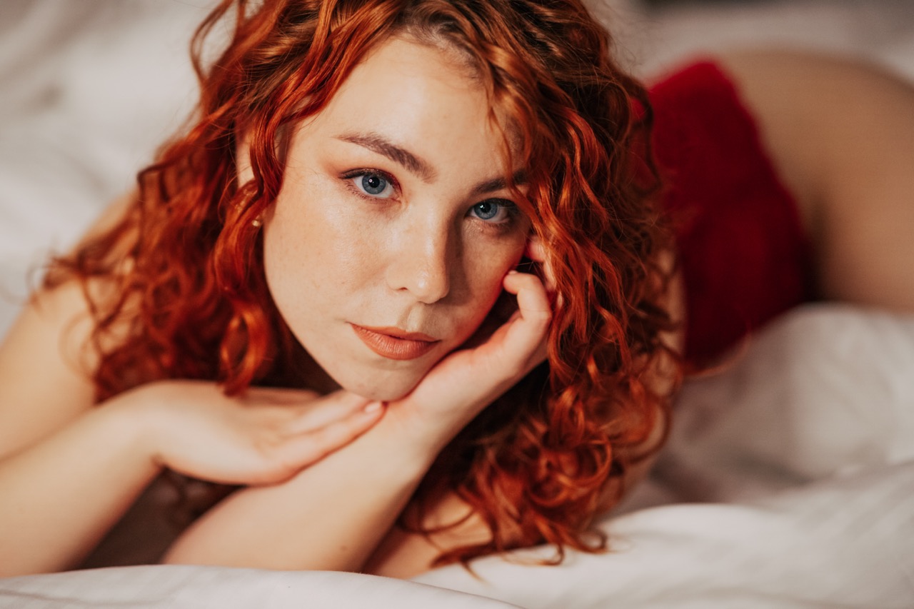 Portret kobiety z rudymi kręconymi włosami i niebieskimi oczami, leżącej na białej pościeli, z delikatnym makijażem i dłońmi pod brodą, patrzącej w obiektyw.