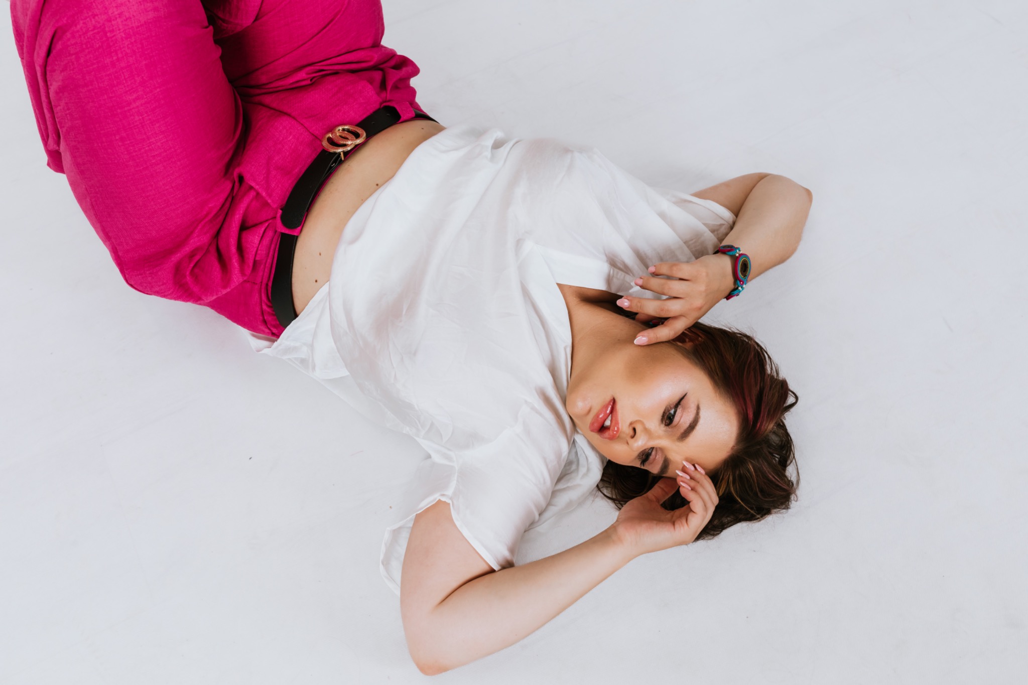 Kobieta leżąca na białym tle, ubrana w różowe spodnie i białą koszulę, z delikatnym makijażem i zegarkiem na ręce.