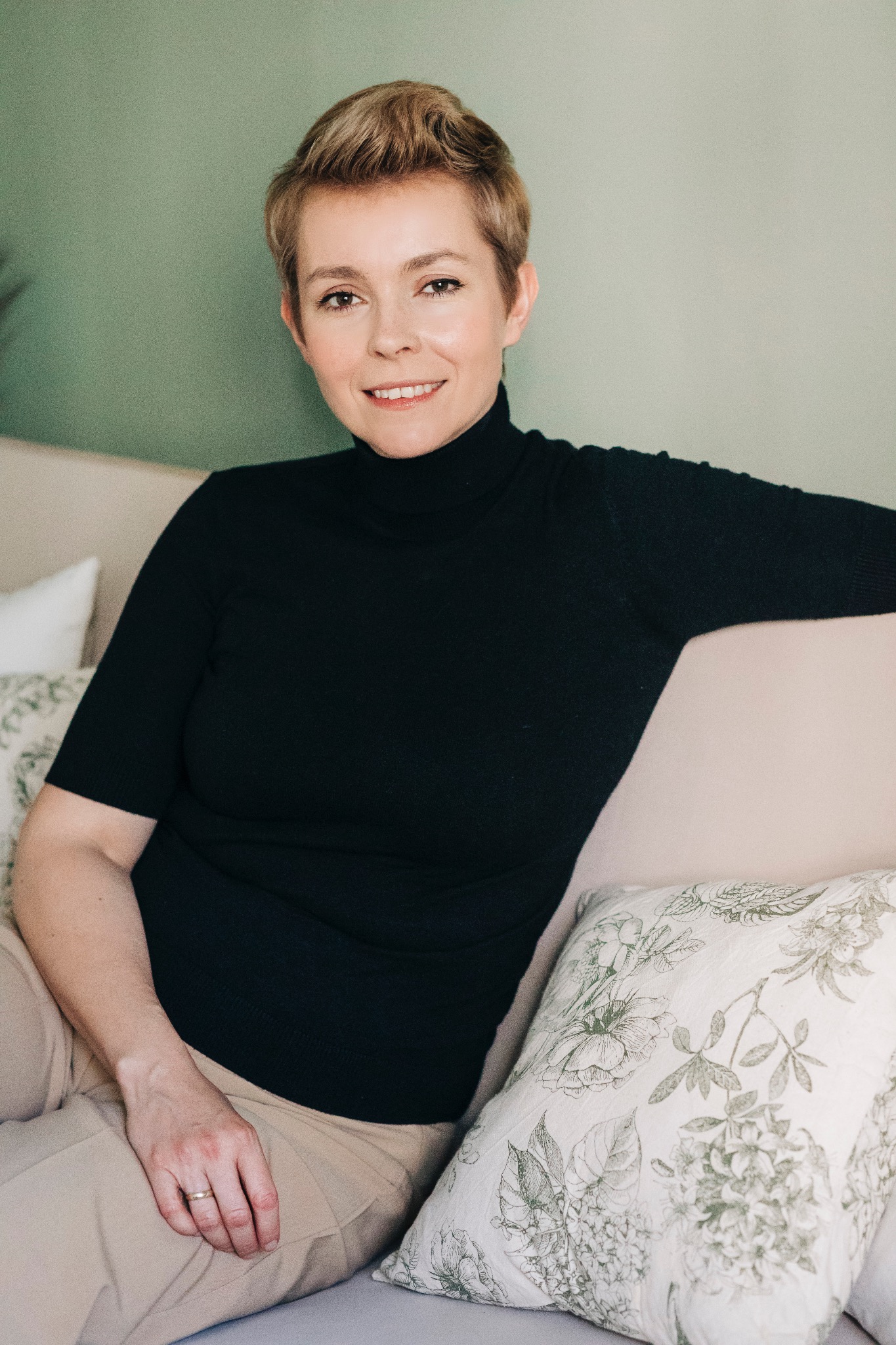 Portret uśmiechniętej kobiety z krótkimi blond włosami w czarnym golfie, siedzącej na jasnej sofie z poduszkami w kwiatowe wzory na tle zielonej ściany.