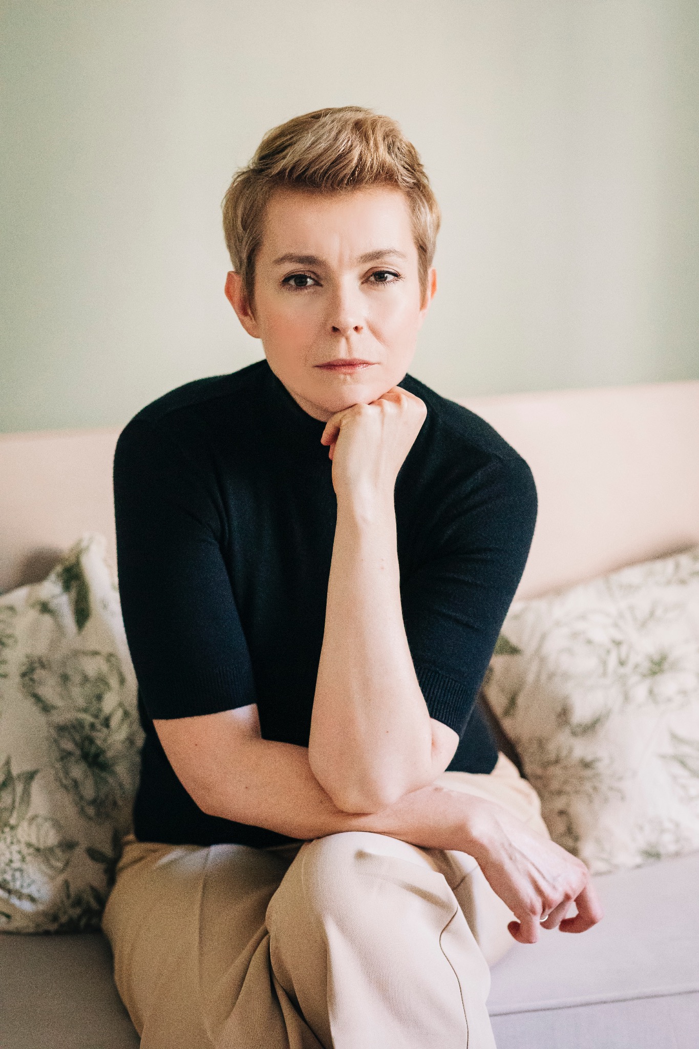 Portret kobiety z krótkimi blond włosami, ubrana w czarny golf, opierająca podbródek na dłoni, siedząca na sofie z poduszką w kwiaty, poważny wyraz twarzy.