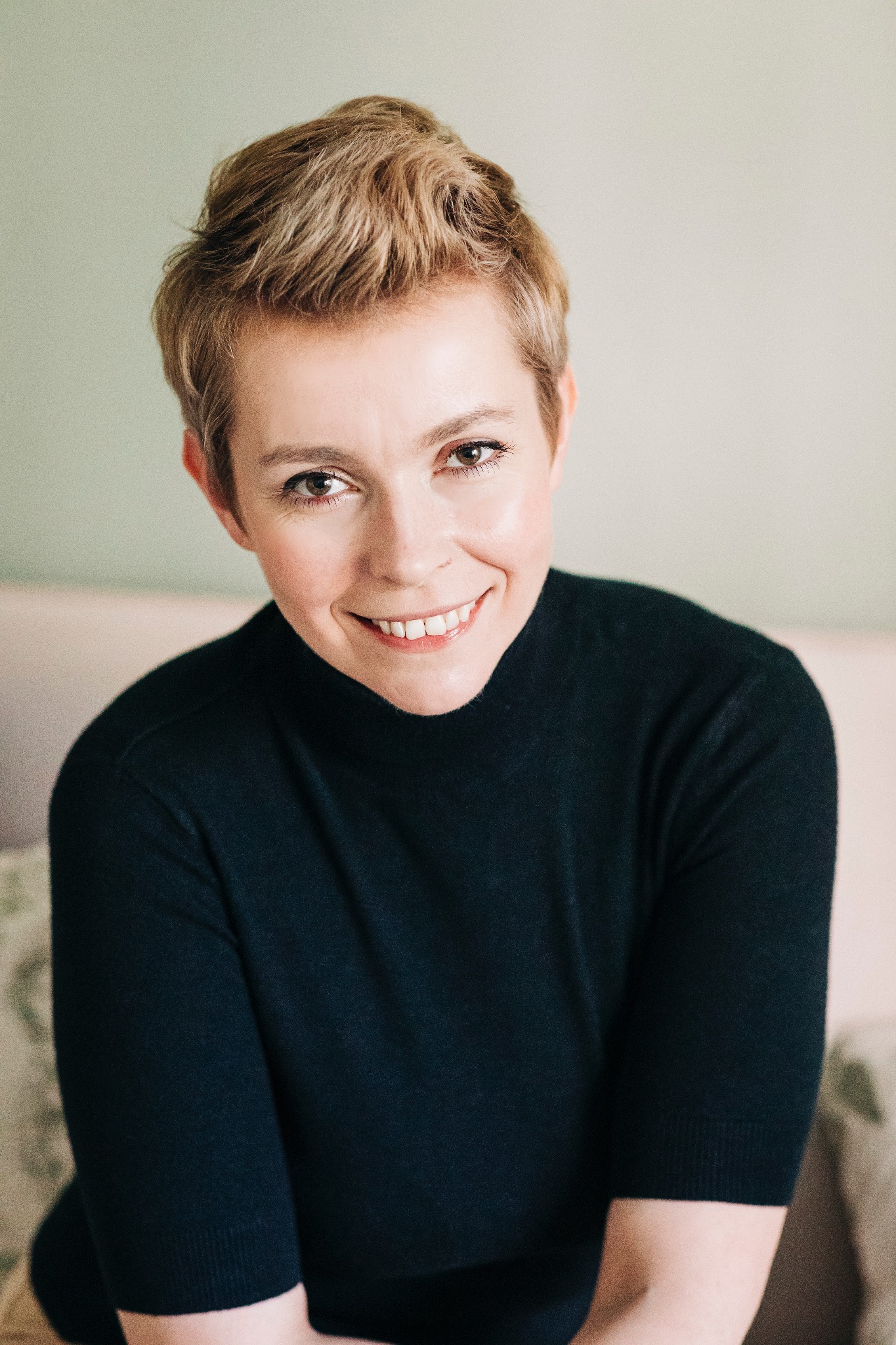 Portret uśmiechniętej kobiety z krótkimi blond włosami, ubrana w czarny golf, z delikatnym makijażem i jasnym tłem.