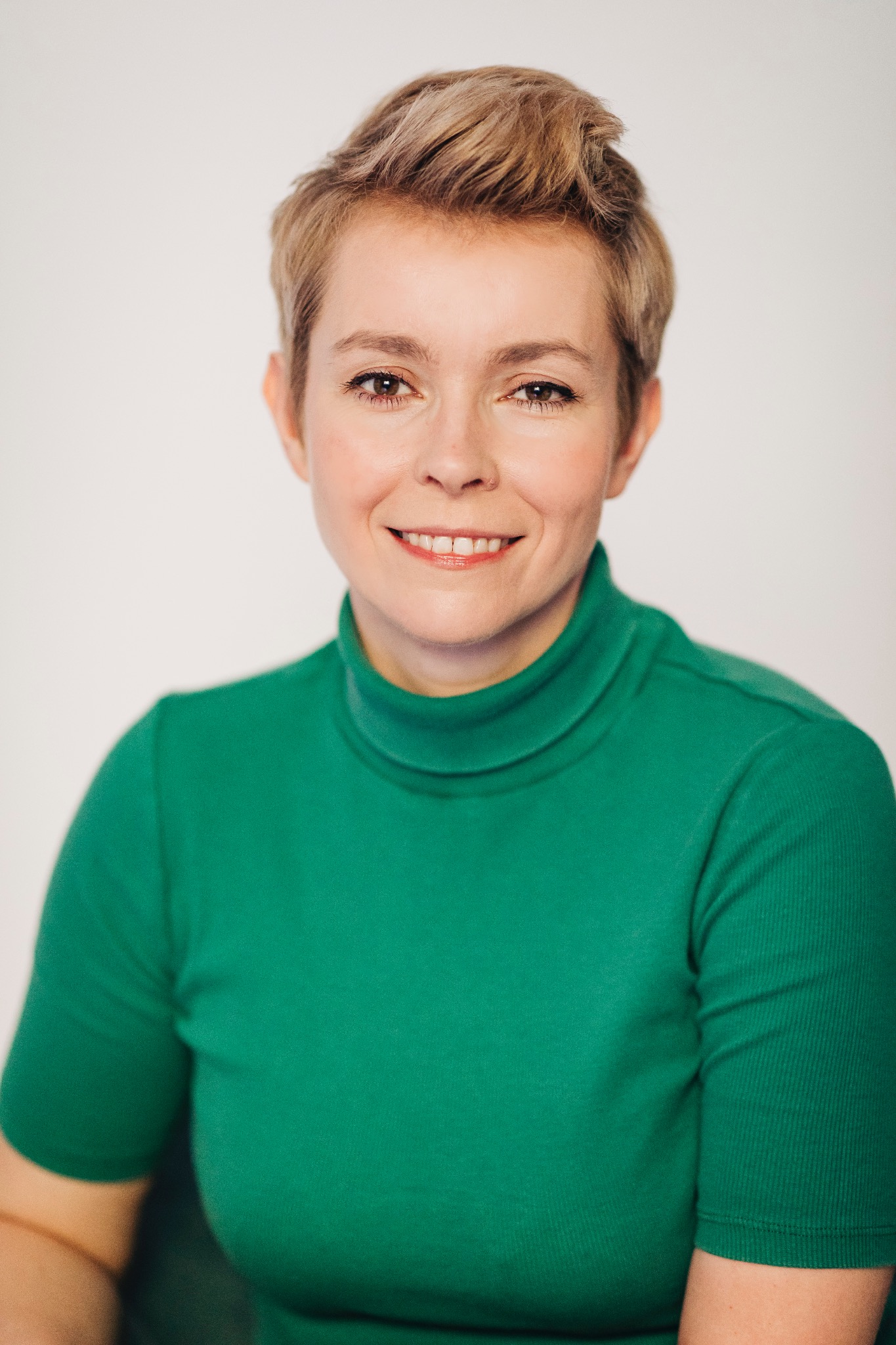 Portret uśmiechniętej kobiety z krótkimi blond włosami, ubrana w zielony golf na jasnym tle.