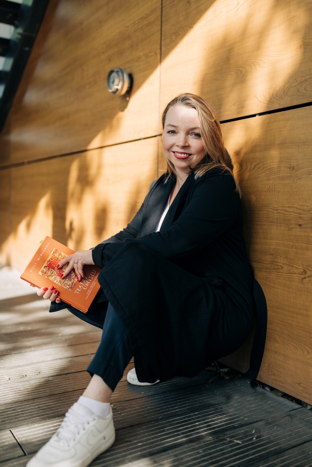 Uśmiechnięta blondynka w czarnym żakiecie, siedząca na drewnianej podłodze przy ścianie z okładziną, trzymająca pomarańczową książkę 'Teatr Umysłu' z czerwoną grafiką.