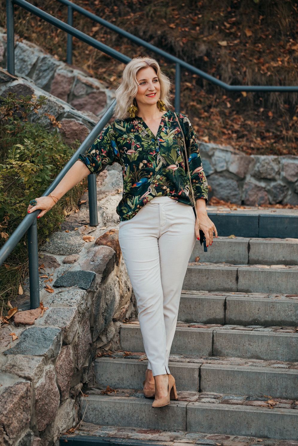 Kobieta w bluzce z motywem tropikalnym i beżowych spodniach pozuje na kamiennych schodach z metalową barierką, uśmiechając się w bok, z zielonymi kolczykami i pierścionkiem.