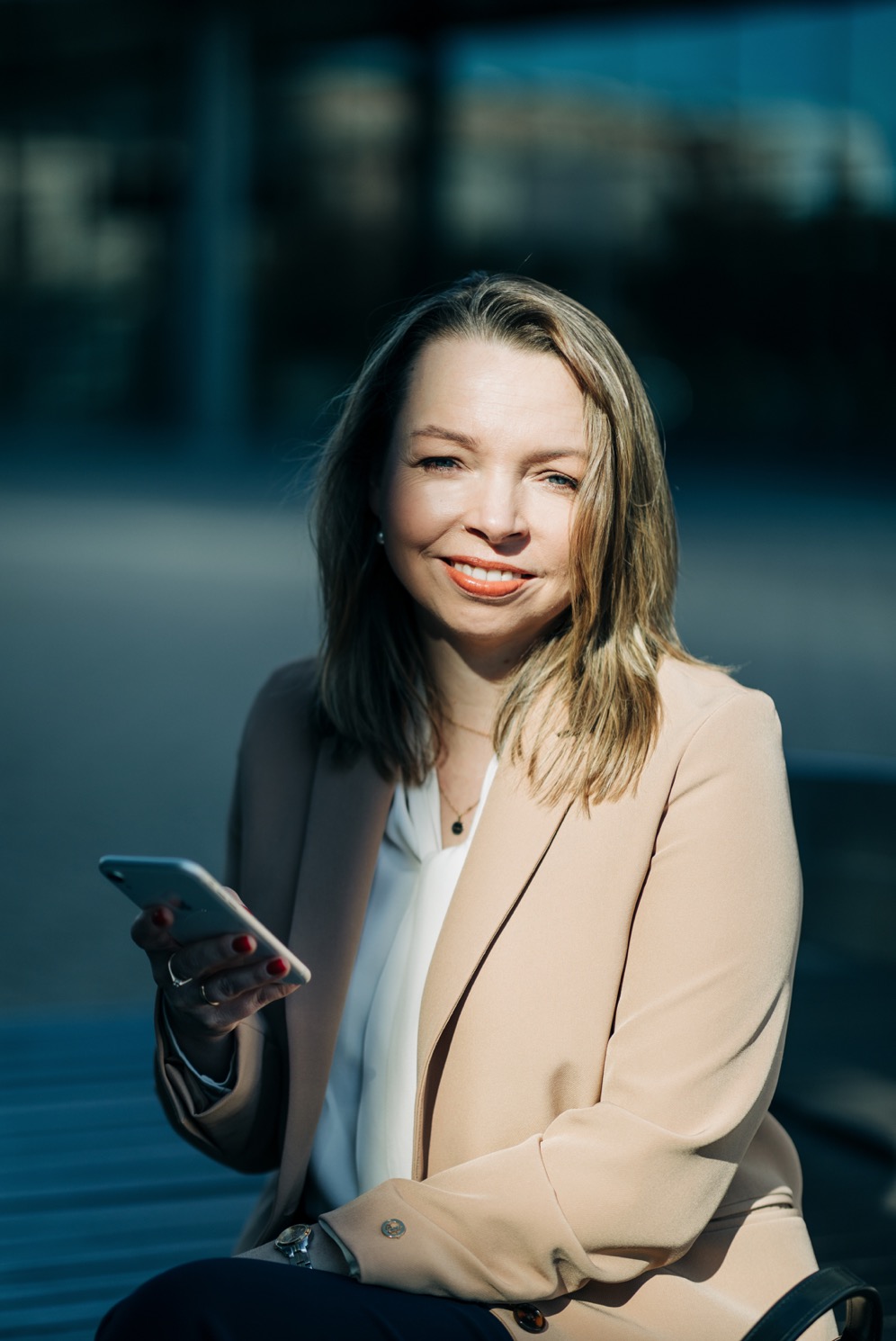 Portret uśmiechniętej kobiety w beżowym żakiecie, trzymającej smartfon na tle miejskiego pejzażu.