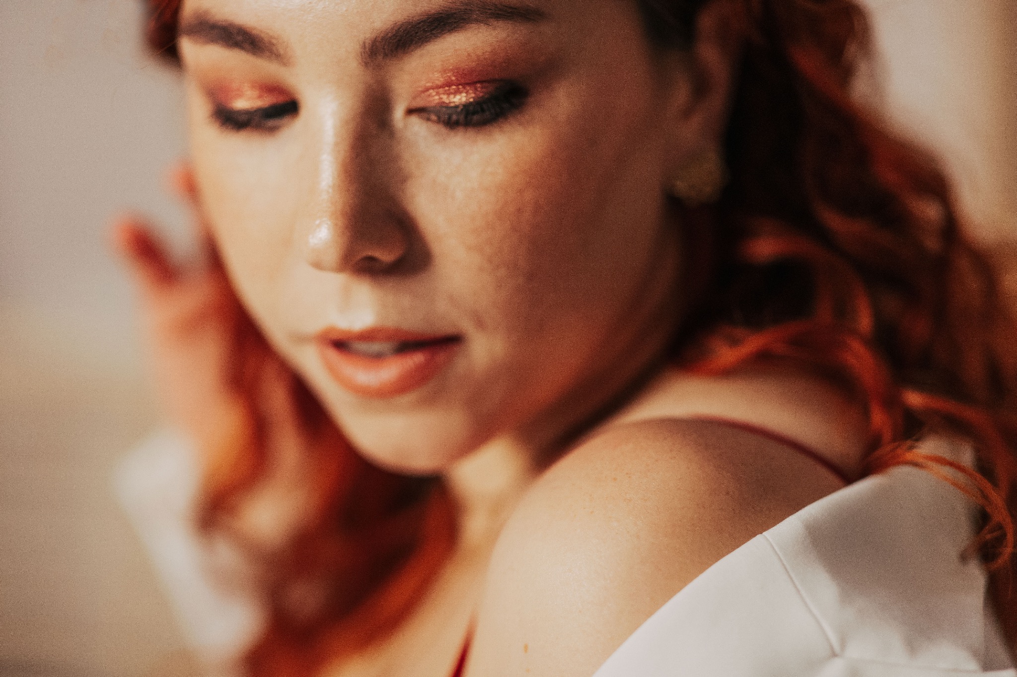 Portret kobiety z rudymi włosami, z delikatnym makijażem oczu w odcieniach brązu i złota, ubrana w białą koszulę, zamyślona poza.