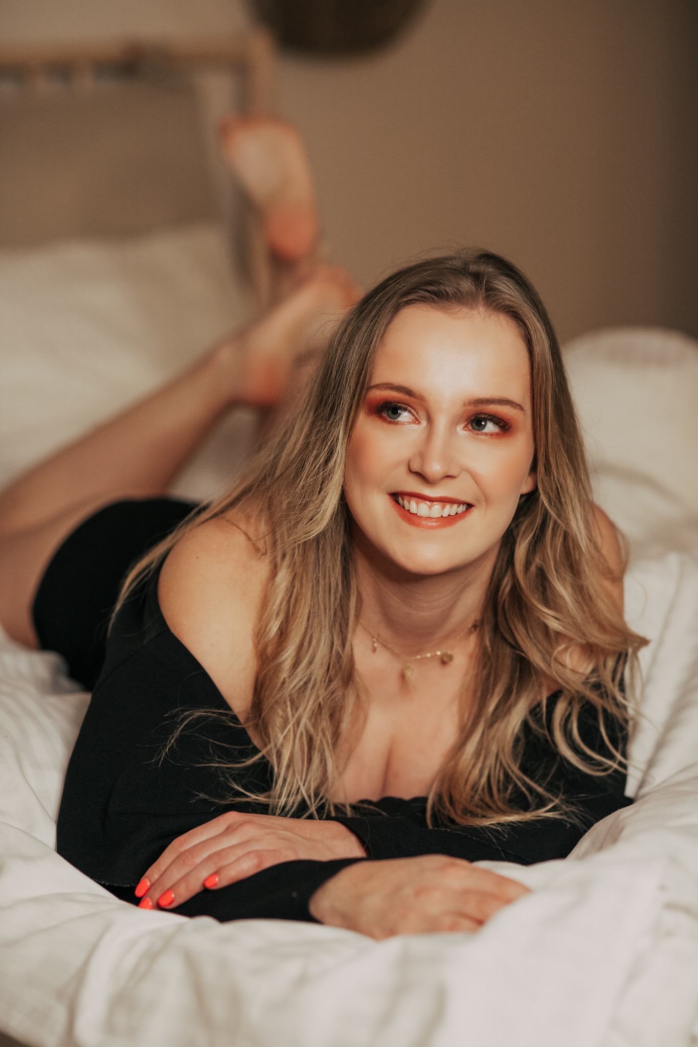 Uśmiechnięta blondynka z pomarańczowym makijażem leży na brzuchu na białej pościeli, paznokcie pomalowane na neonowy pomarańcz, delikatna złota biżuteria.