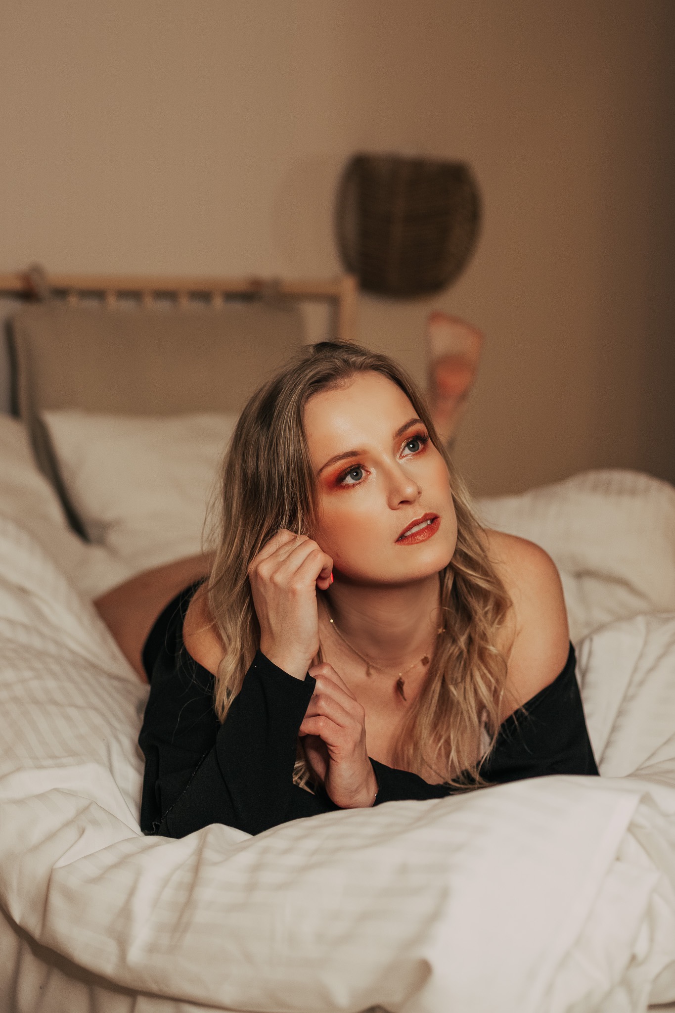 Portret kobiety z blond włosami leżącej na łóżku, z intensywnym makijażem oczu w odcieniach pomarańczy i złota, patrzącej w górę.