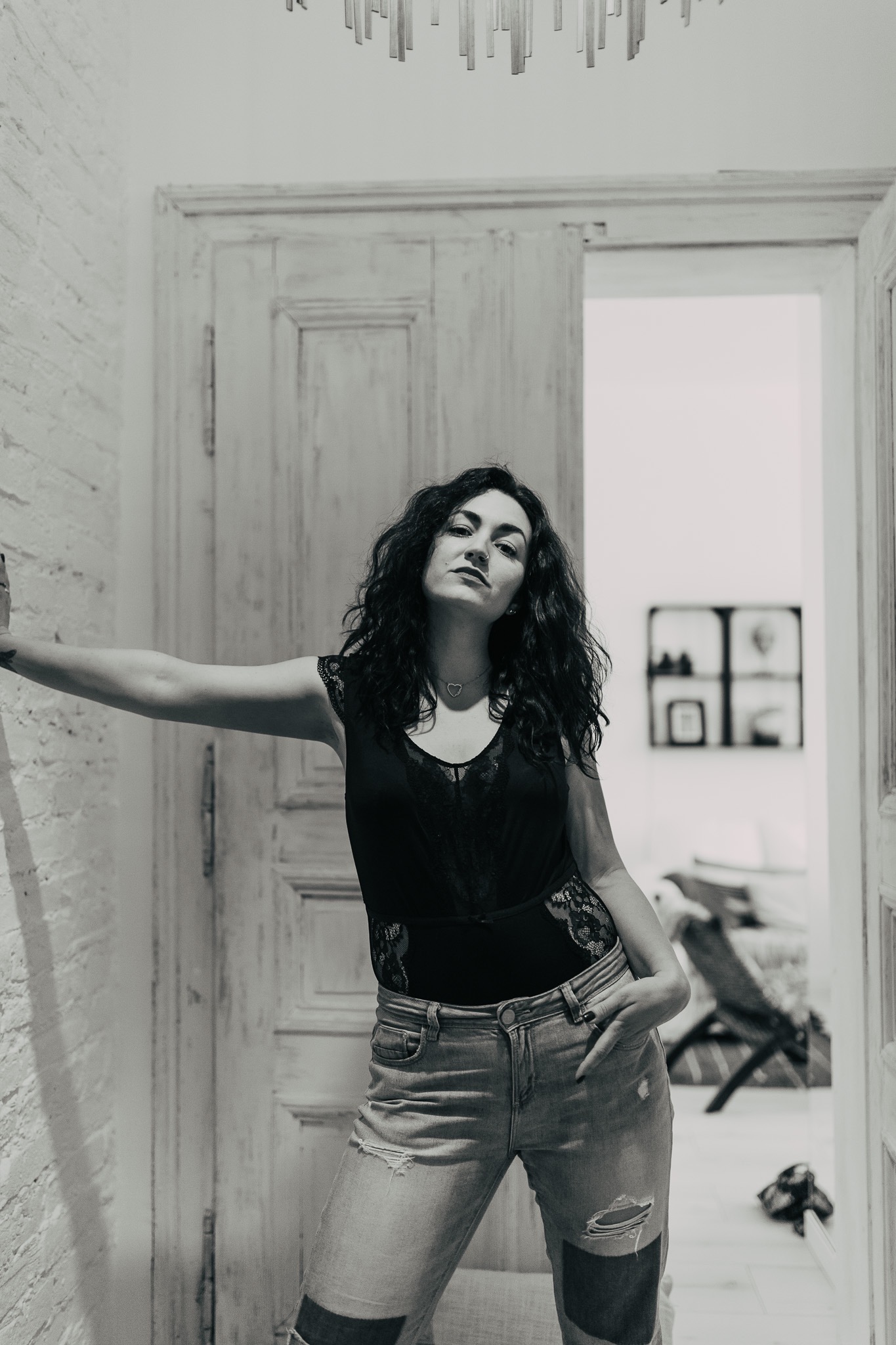 Czarno-białe zdjęcie kobiety w koronkowym body i podartych jeansach, opierającej się o ścianę przy otwartych, drewnianych drzwiach.