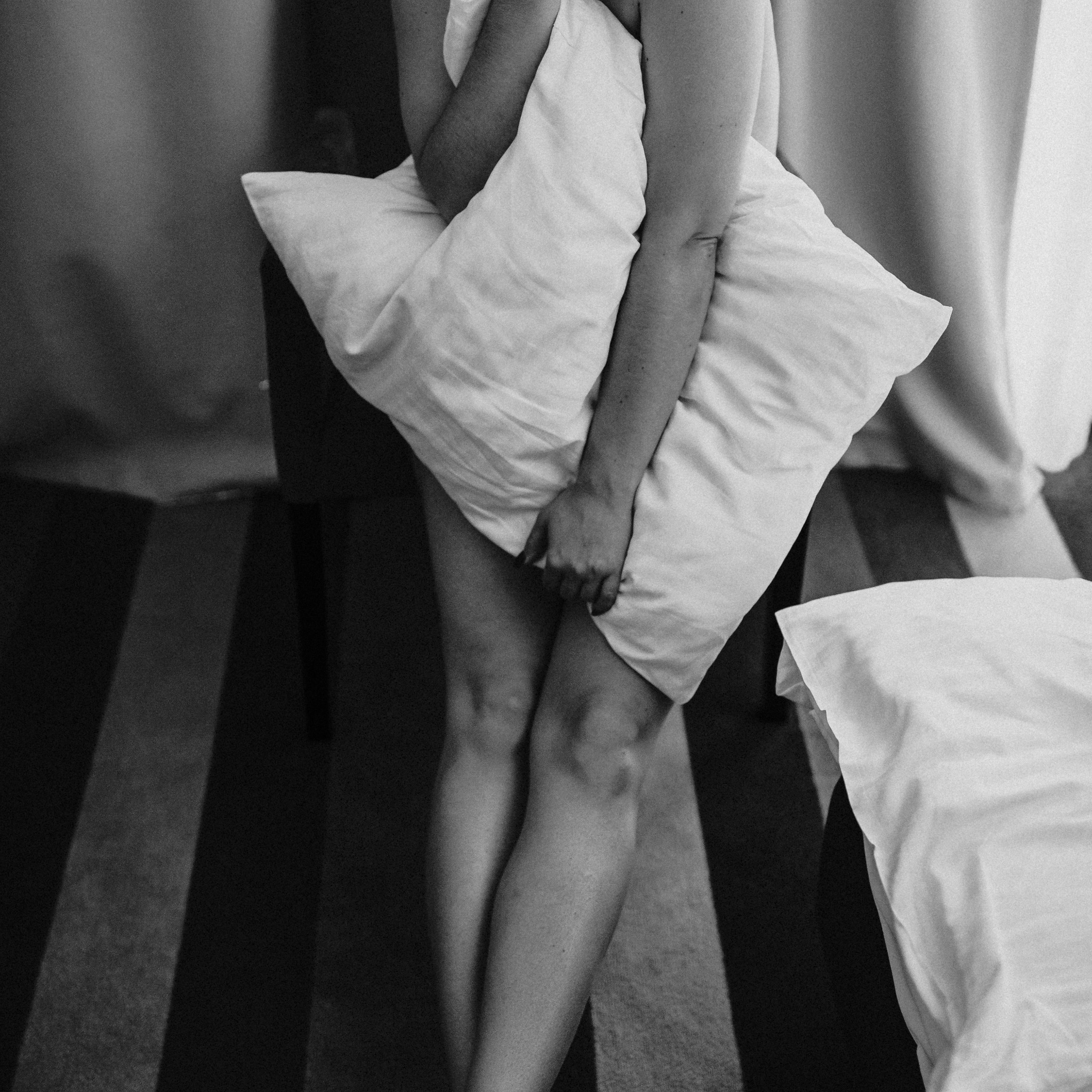 Czarno-białe zdjęcie kobiety trzymającej białą poduszkę, widoczne nogi i ręce, minimalistyczne tło z zasłonami i dywanem w paski.