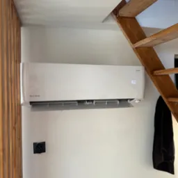 Biały klimatyzator Rotenso zamontowany na ścianie w pomieszczeniu z drewnianymi schodami i dekoracyjną ścianką z listewek.