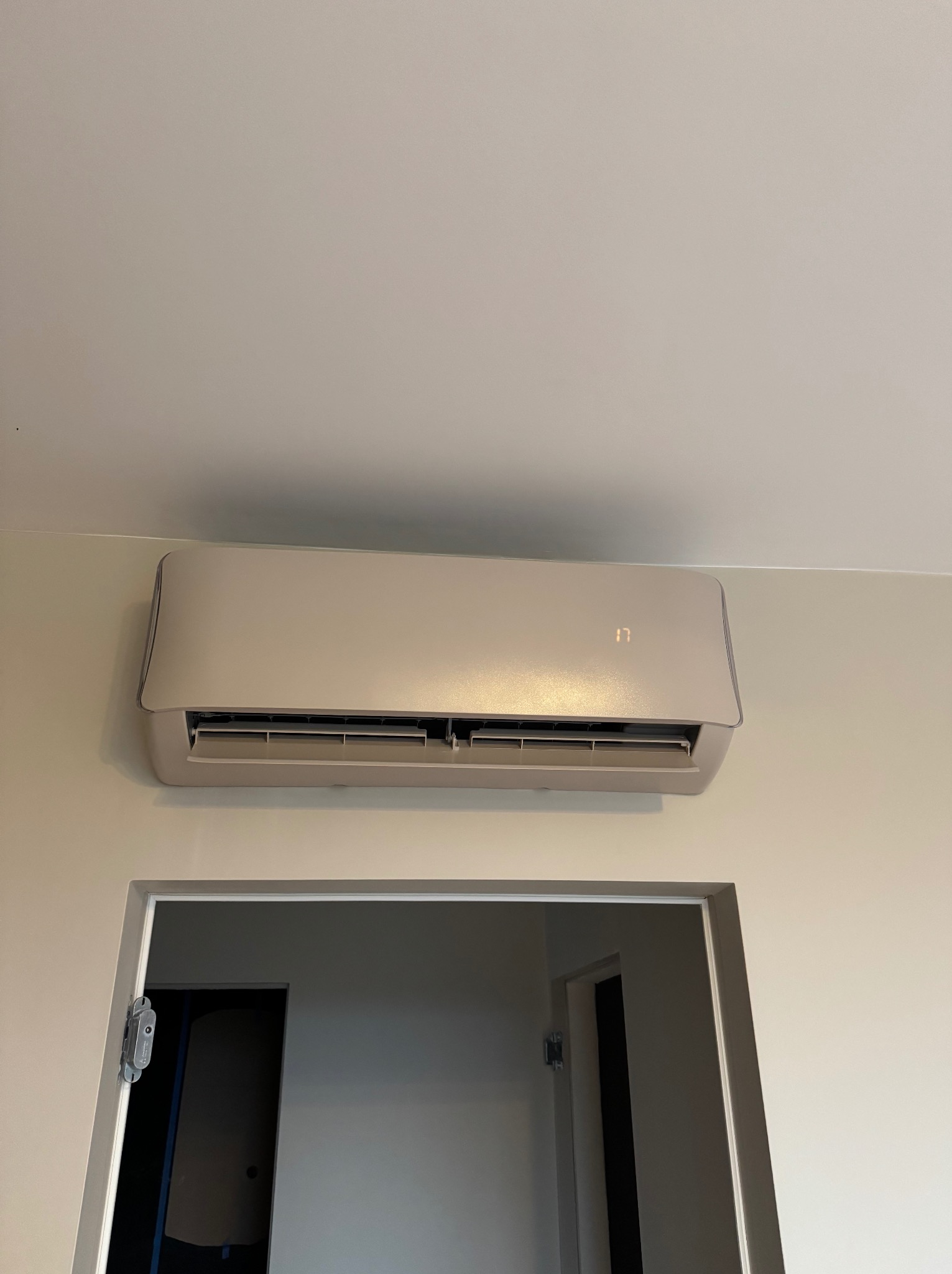 Beżowy klimatyzator ścienny z wyświetlaczem temperatury, zamontowany nad otwartymi drzwiami w jasnym pomieszczeniu.