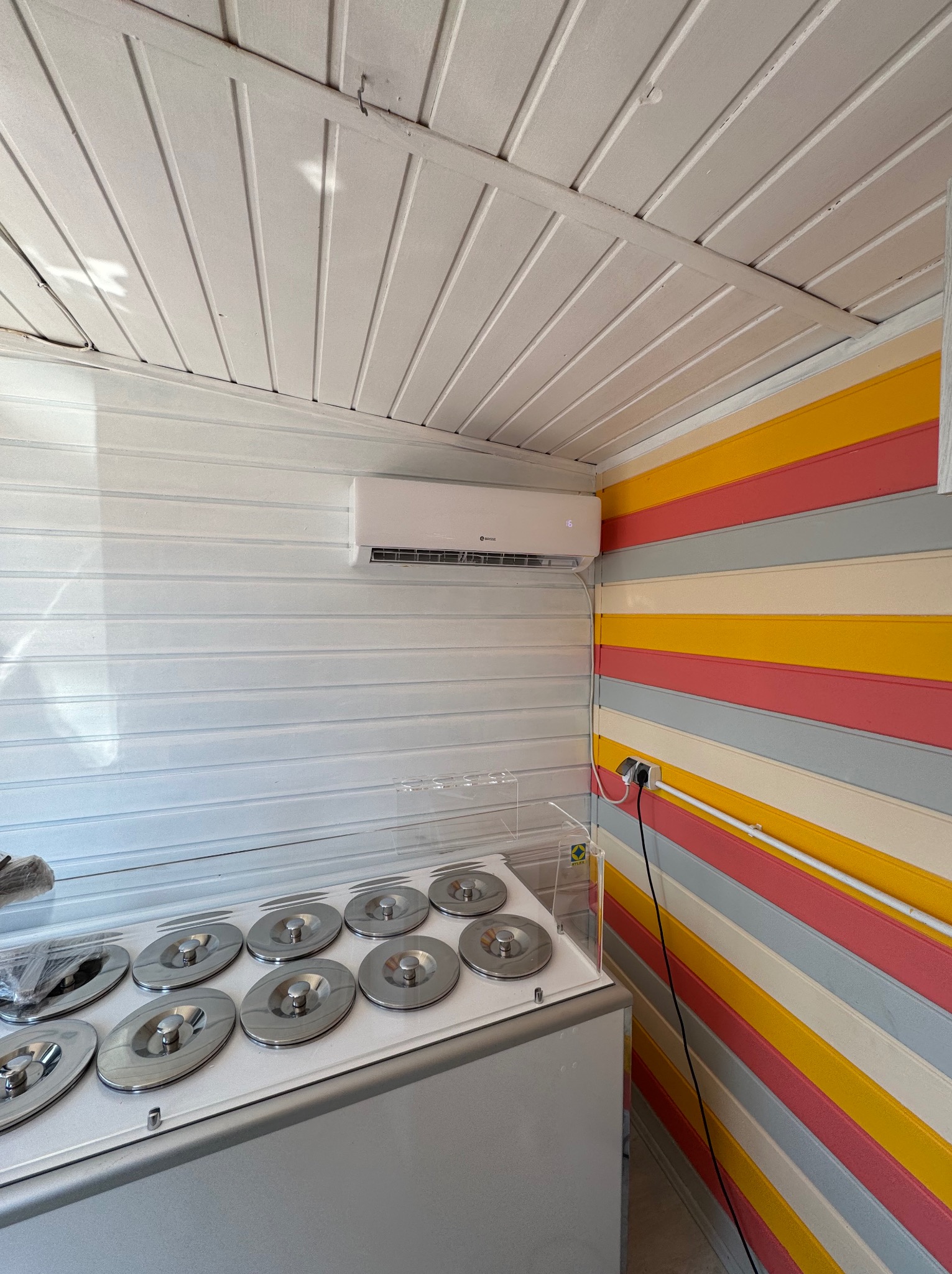 Wnętrze lokalu z zainstalowanym klimatyzatorem na ścianie z białych paneli, obok ściana w poziome pasy: żółty, różowy, szary i kremowy, pod spodem zamrażarka do lodów z metalowymi pokrywami.