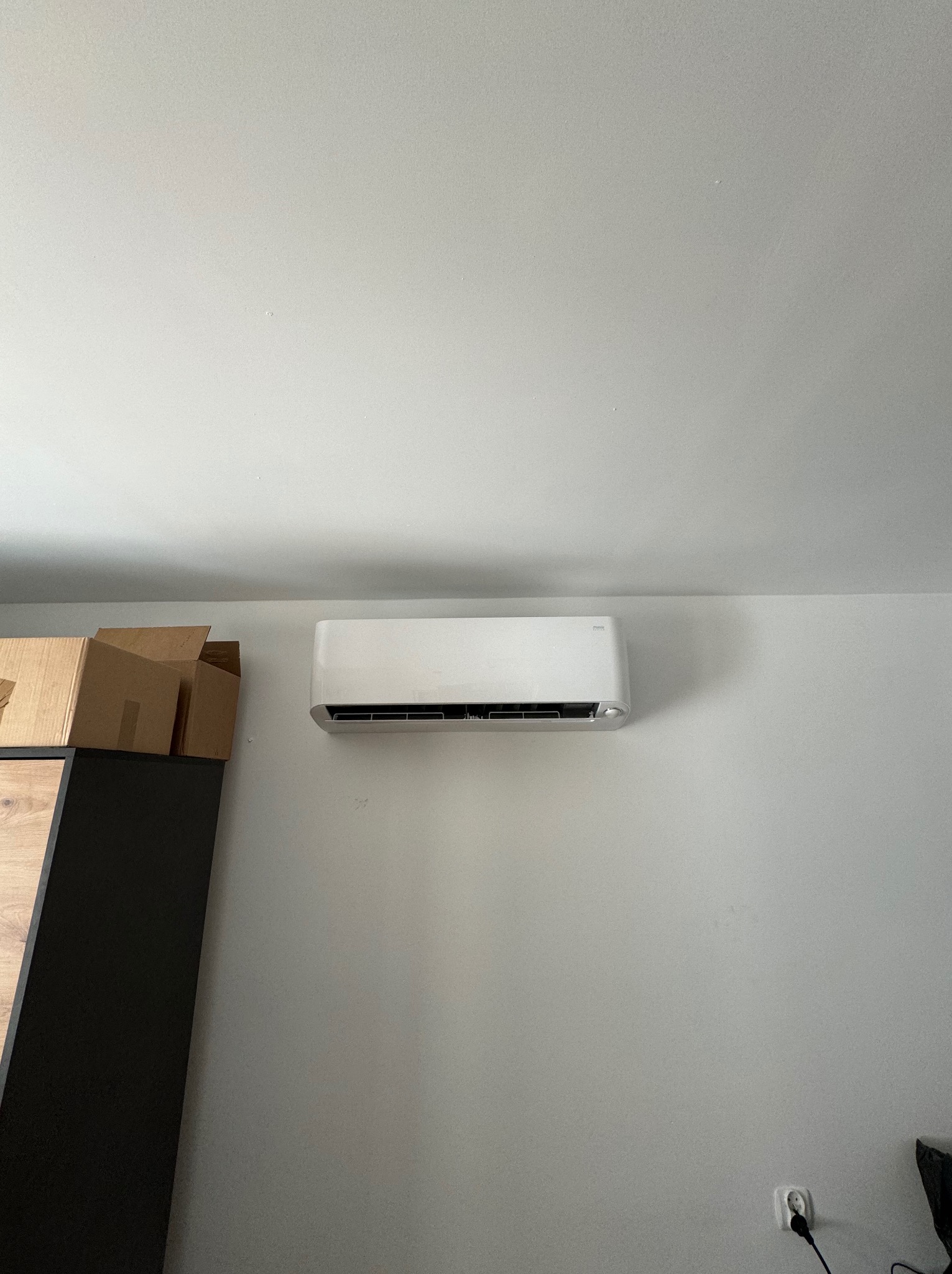 Biały klimatyzator zamontowany na ścianie w pokoju, widoczny fragment szafki z kartonami po lewej stronie i gniazdko elektryczne na dole ściany.