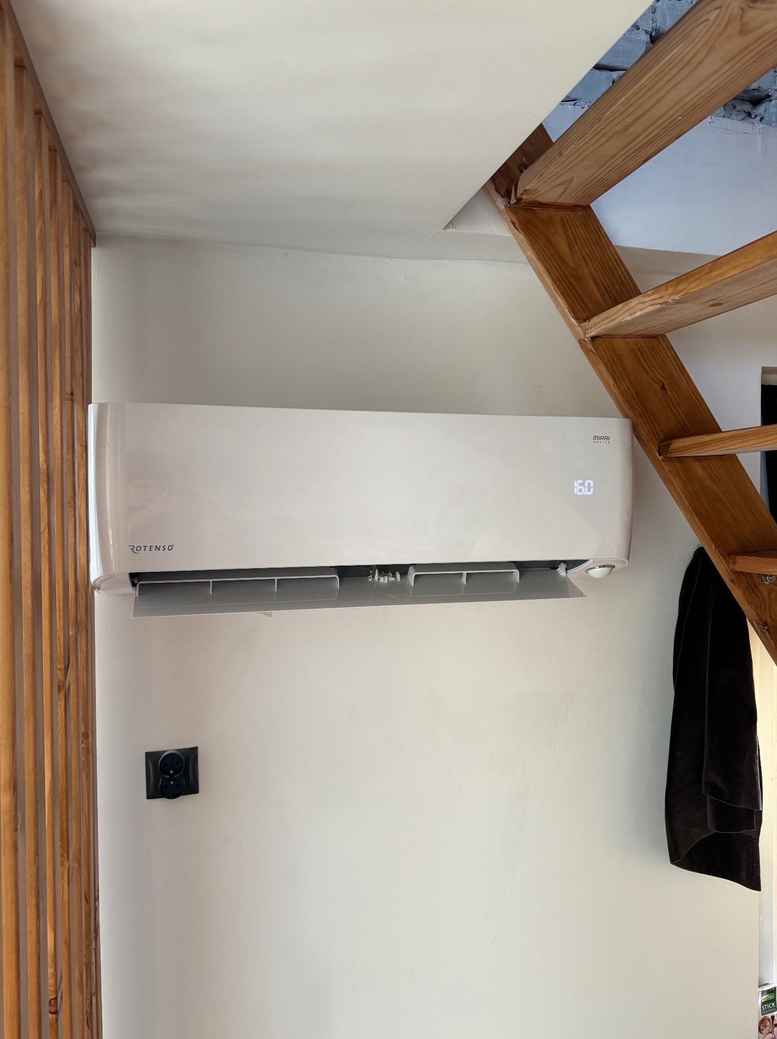 Biały klimatyzator Rotenso zamontowany na ścianie w pomieszczeniu z drewnianymi schodami i dekoracyjną ścianką z listewek.