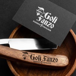 Blank Sp. z o.o. - Czarna wizytówka oraz brzytwa z drewnianą rączką, obie z logo 'Goli Fanzo Barber Fryzjer', ułożone w drewnianej miseczce na czarnym tle.