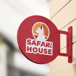 Blank Sp. z o.o. - Okrągły, czerwony szyld z białym napisem SAFARI HOUSE i sylwetką psa w pomarańczowym okręgu, zamocowany na beżowej ścianie.