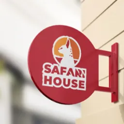 Okrągły, czerwony szyld z białym napisem SAFARI HOUSE i sylwetką psa w pomarańczowym okręgu, zamocowany na beżowej ścianie.