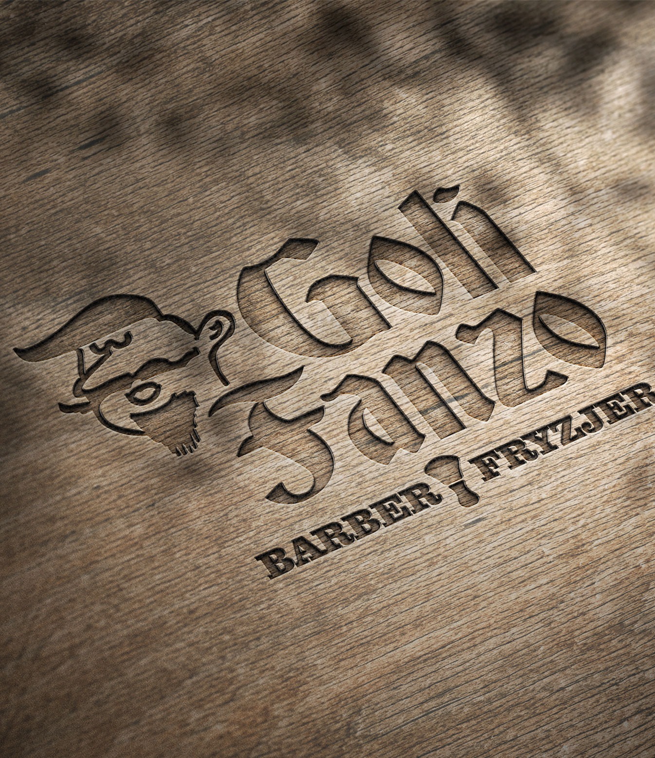 Wygrawerowane logo barber shopu 'Goli Fanze Barber Fryzjer' z wizerunkiem brodatego mężczyzny w kapeluszu na drewnianej desce z widoczną strukturą drewna.