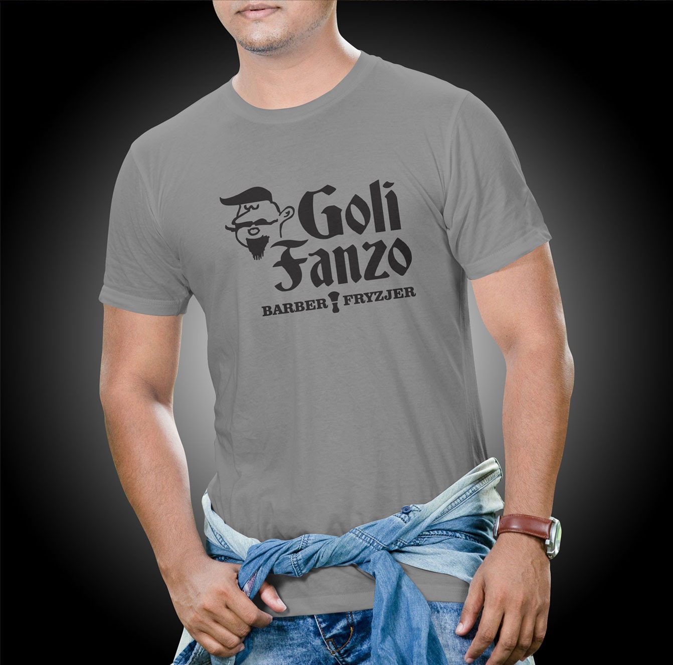 Mężczyzna w szarej koszulce z czarnym nadrukiem 'Goli Fanzo BARBER FRYZJER' i grafiką brodatego mężczyzny, jeansowa koszula zawiązana w pasie, na nadgarstku brązowy zegarek.