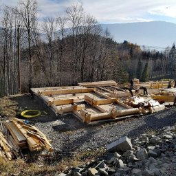 Wojciech Kędryna Usługi Budowlane-Dekarskie - Budowa domu z bali: fundamenty i pierwsze elementy konstrukcji, widoczny dźwig, góry w tle.