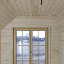 Wojciech Kędryna Usługi Budowlane-Dekarskie - Wnętrze poddasza wykończonego jasnym drewnem, z oknem dwuskrzydłowym z widokiem na krajobraz, widoczne przewody elektryczne.