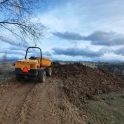 Żółta wywrotka budowlana Mecalac na szczycie pryzmy ziemi, widoczne ślady opon, w tle wiejski krajobraz z górami i zachmurzonym niebem.