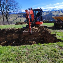 Wojciech Kędryna Usługi Budowlane-Dekarskie - Pomarańczowa mini koparka Kubota w trakcie wykopywania rowu w ziemi, operator w kabinie, wywrotka w tle, widok na zielone wzgórza i ośnieżone szczyty górskie pod błękitnym niebem.