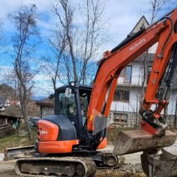 Wojciech Kędryna Usługi Budowlane-Dekarskie - Pomarańczowa mini koparka Kubota U55-4 na nierównym terenie, operator w kabinie, widoczny fragment domu w tle.