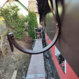 Wojciech Kędryna Usługi Budowlane-Dekarskie - Montaż systemu rynnowego z bliska, widok z rusztowania na brązowe haki rynnowe, narzędzia na parapecie okna, dom w tle.