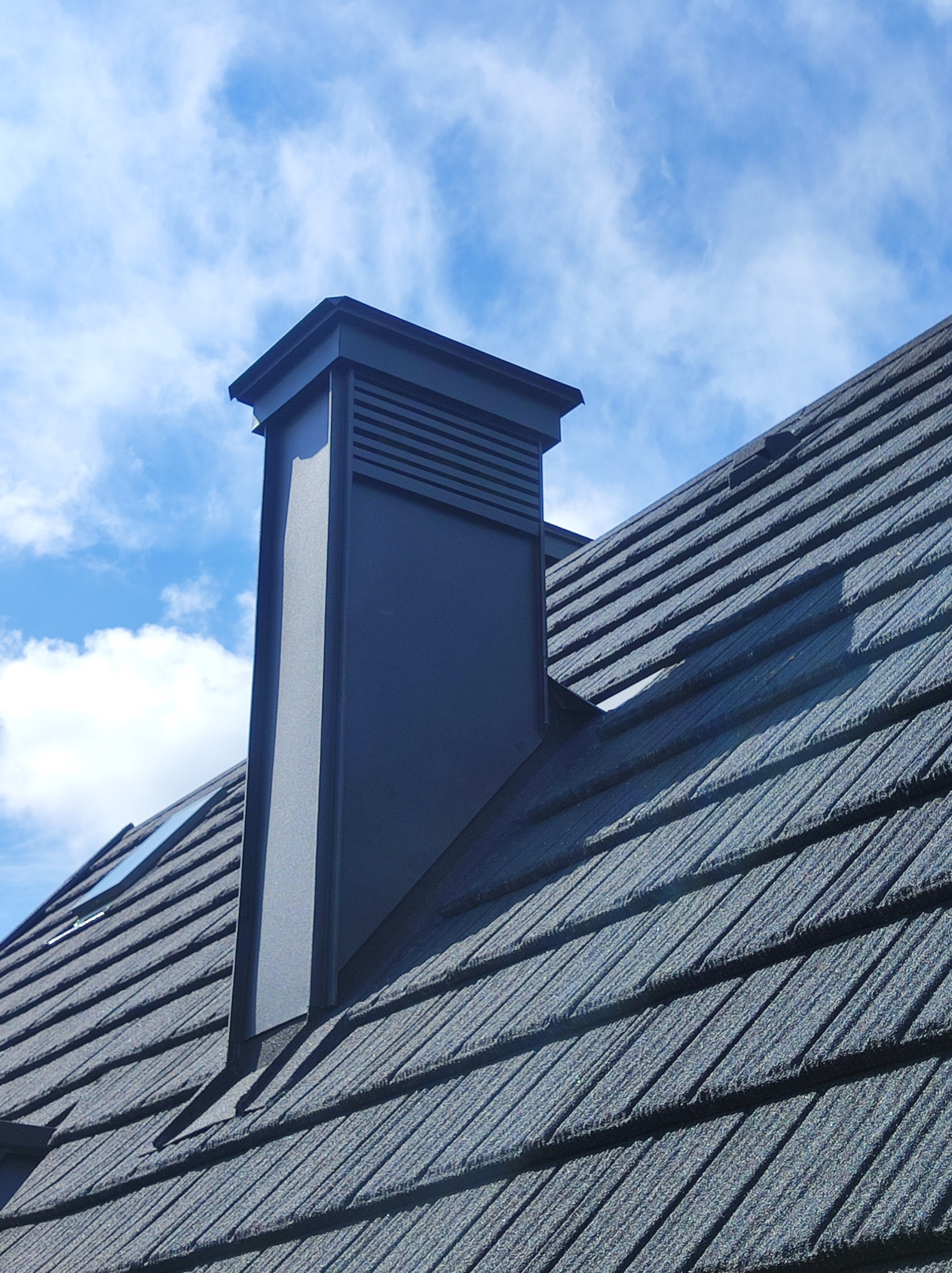 Nowoczesny, grafitowy komin wentylacyjny na dachu krytym ciemnoszarą blachodachówką, na tle błękitnego nieba z lekkimi obłokami.