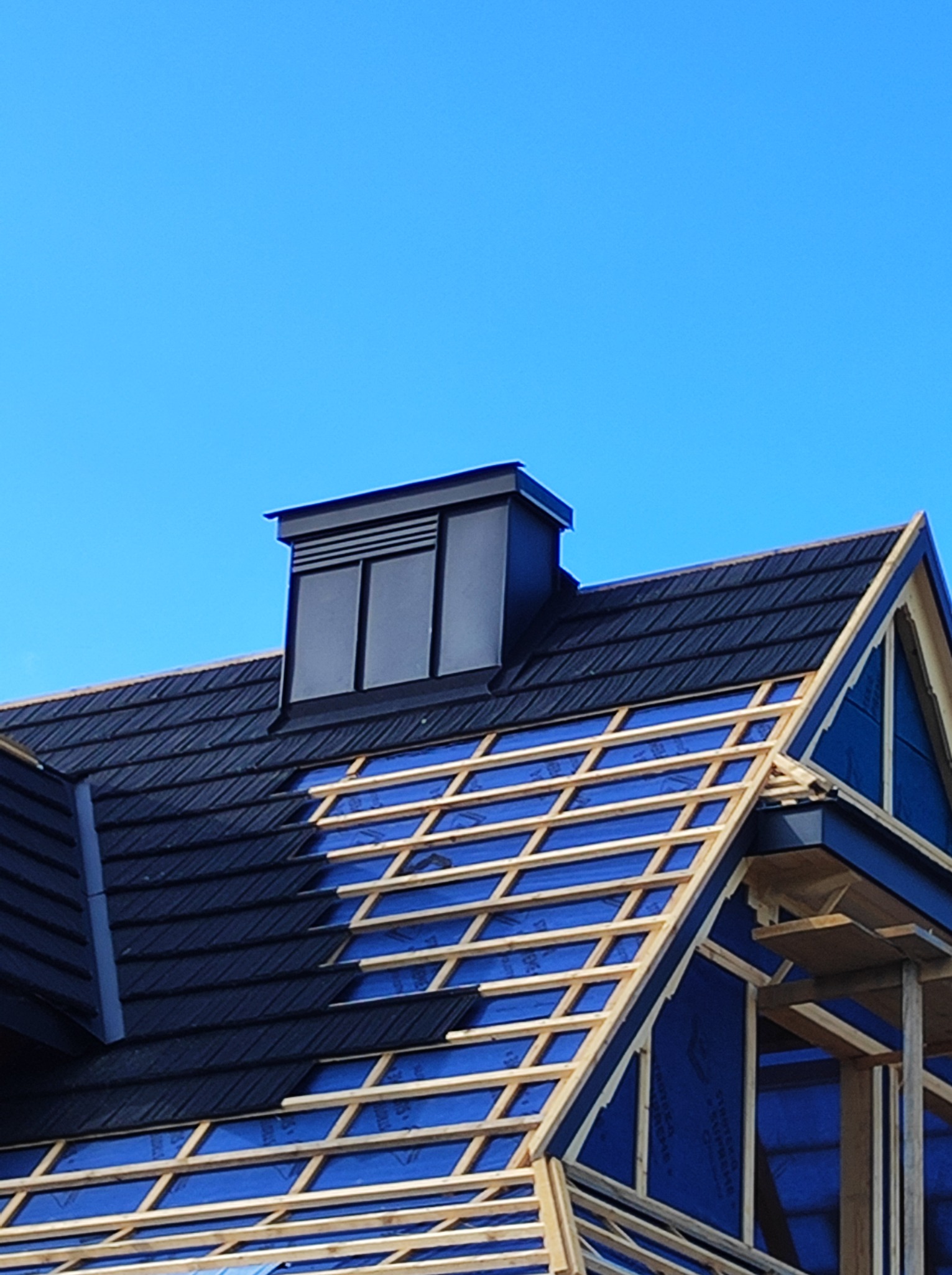 Częściowo pokryty czarną dachówką dach z kominem wentylacyjnym, widoczna więźba dachowa z membraną w kolorze niebieskim.