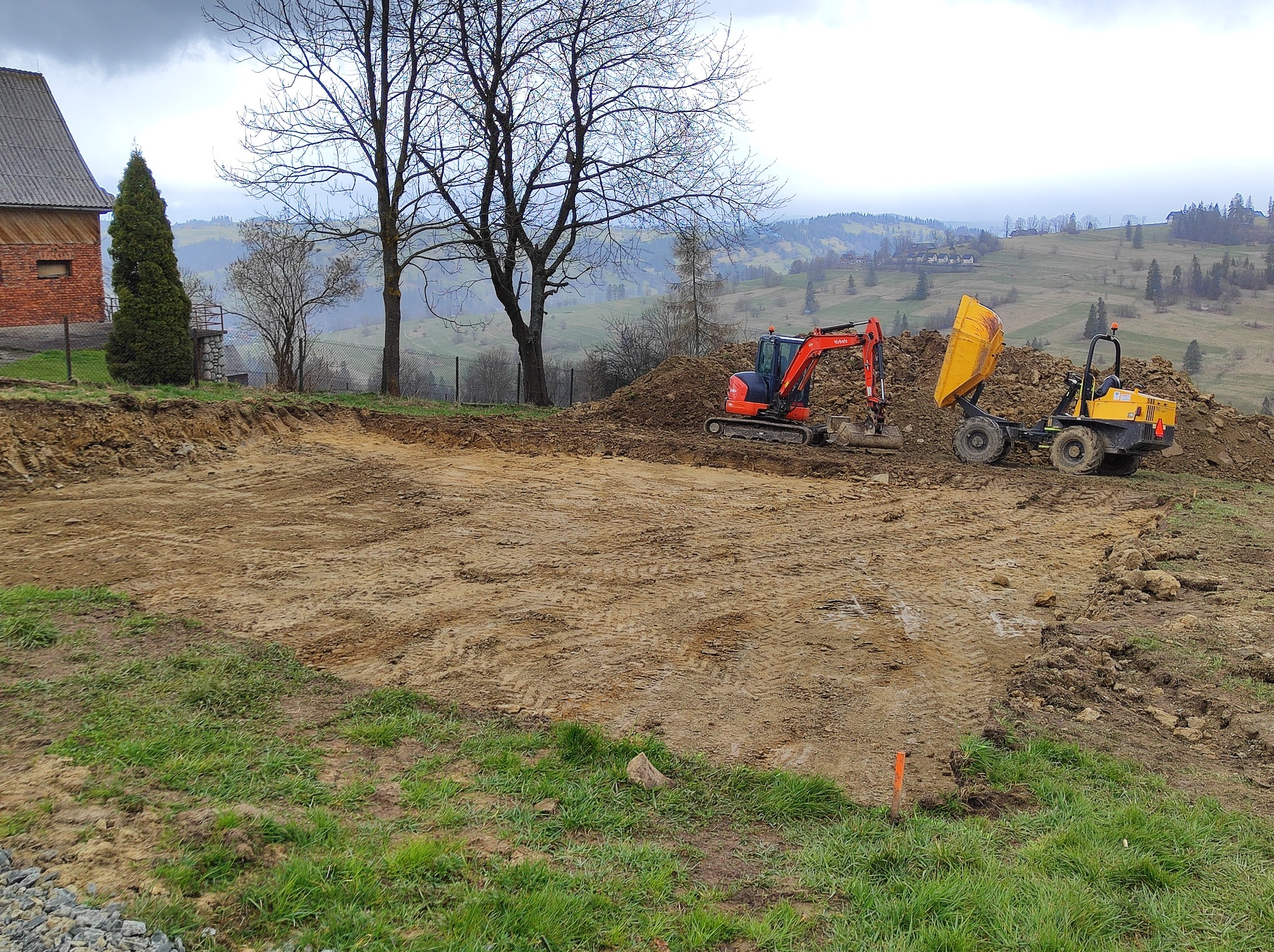 Przygotowanie terenu pod budowę na wzgórzu: wyrównana ziemia, czerwona koparka Kubota i żółta wywrotka na tle pagórkowatego krajobrazu.