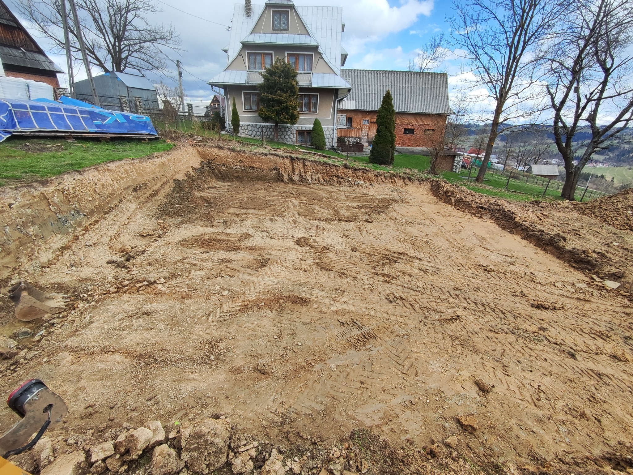 Wykonany wykop pod fundamenty domu jednorodzinnego na działce w górskim krajobrazie, widoczne ślady opon koparki na gliniastej ziemi.