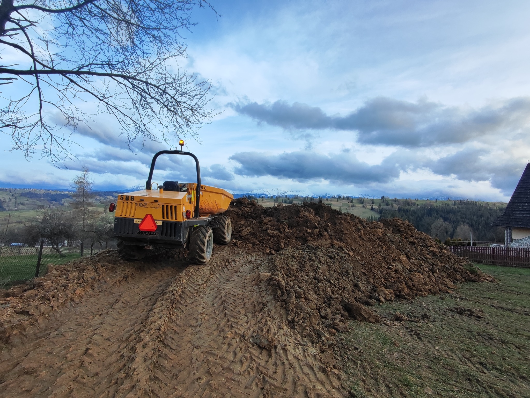 Żółta wywrotka budowlana Mecalac na szczycie pryzmy ziemi, widoczne ślady opon, w tle wiejski krajobraz z górami i zachmurzonym niebem.