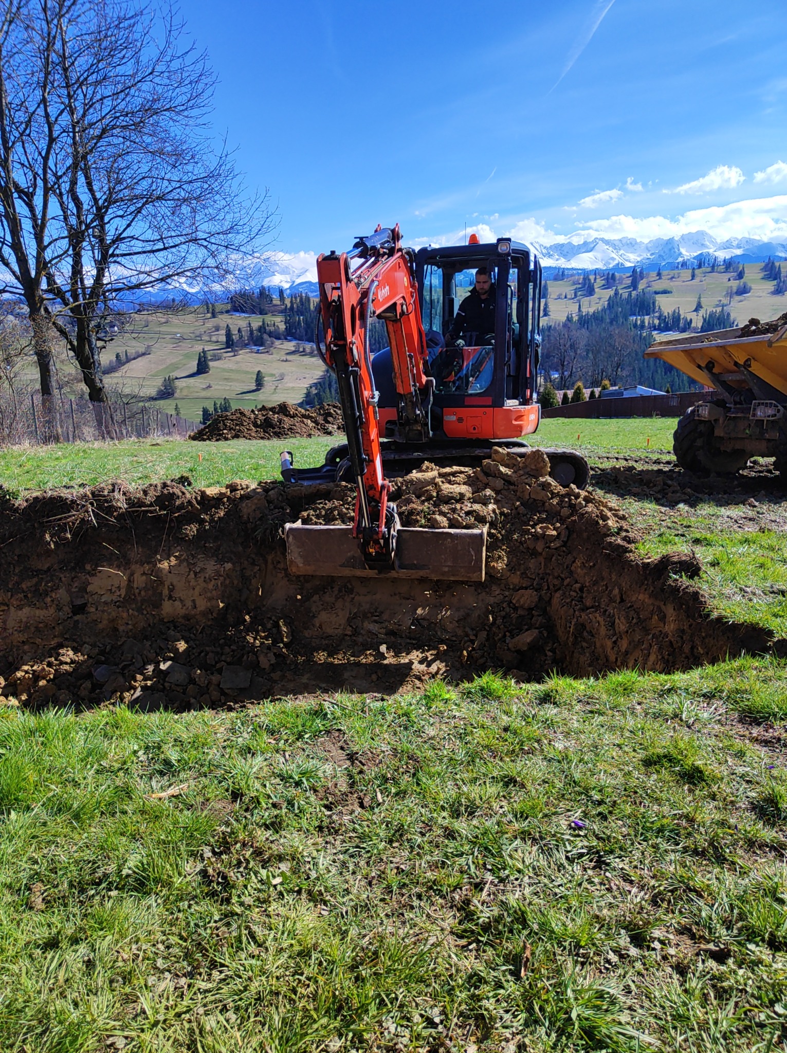 Pomarańczowa mini koparka Kubota w trakcie wykopywania rowu w ziemi, operator w kabinie, wywrotka w tle, widok na zielone wzgórza i ośnieżone szczyty górskie pod błękitnym niebem.