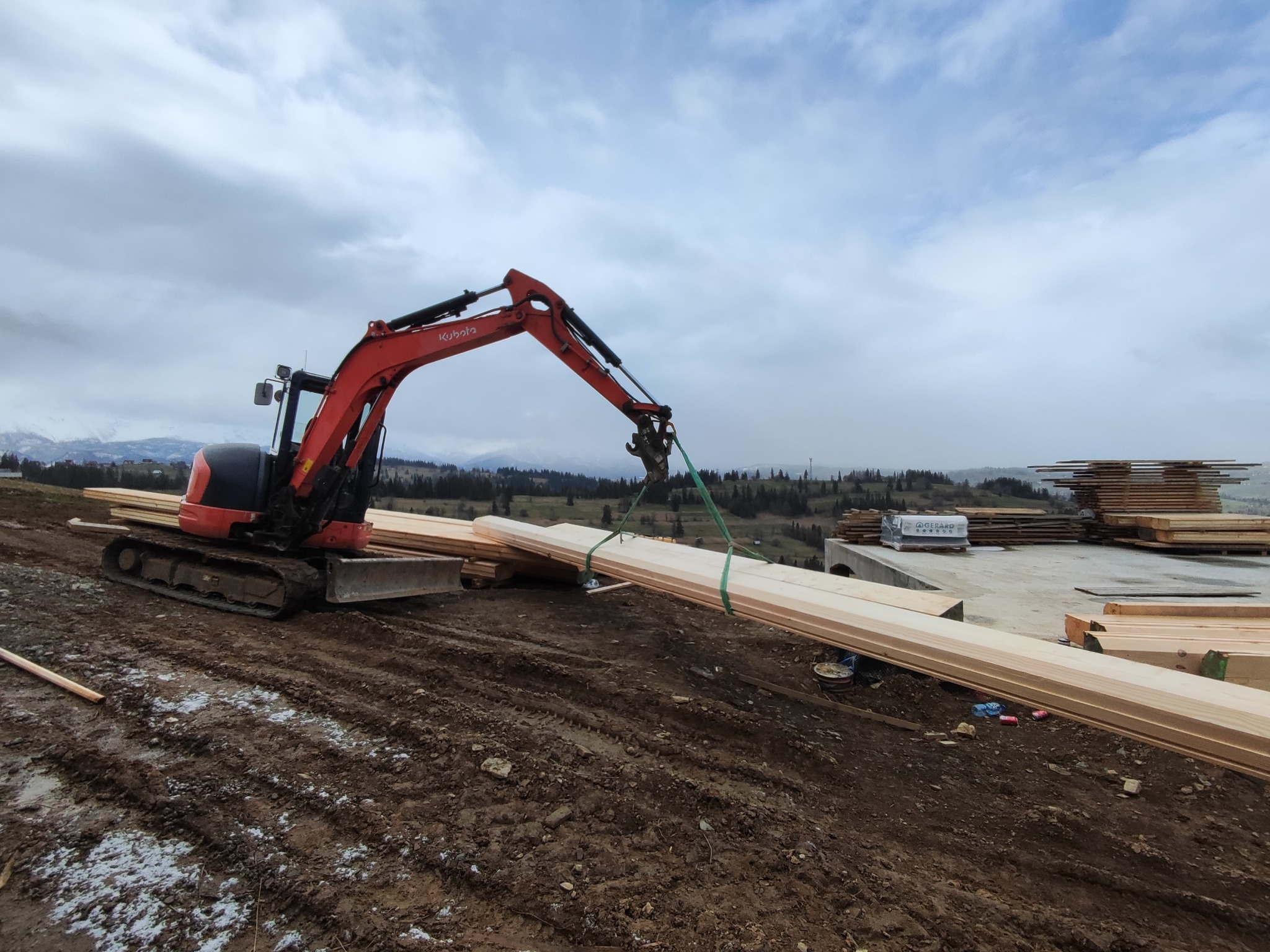 Czerwona mini koparka Kubota podnosi wiązkę jasnego drewna na placu budowy, w tle widoczne góry i pochmurne niebo.