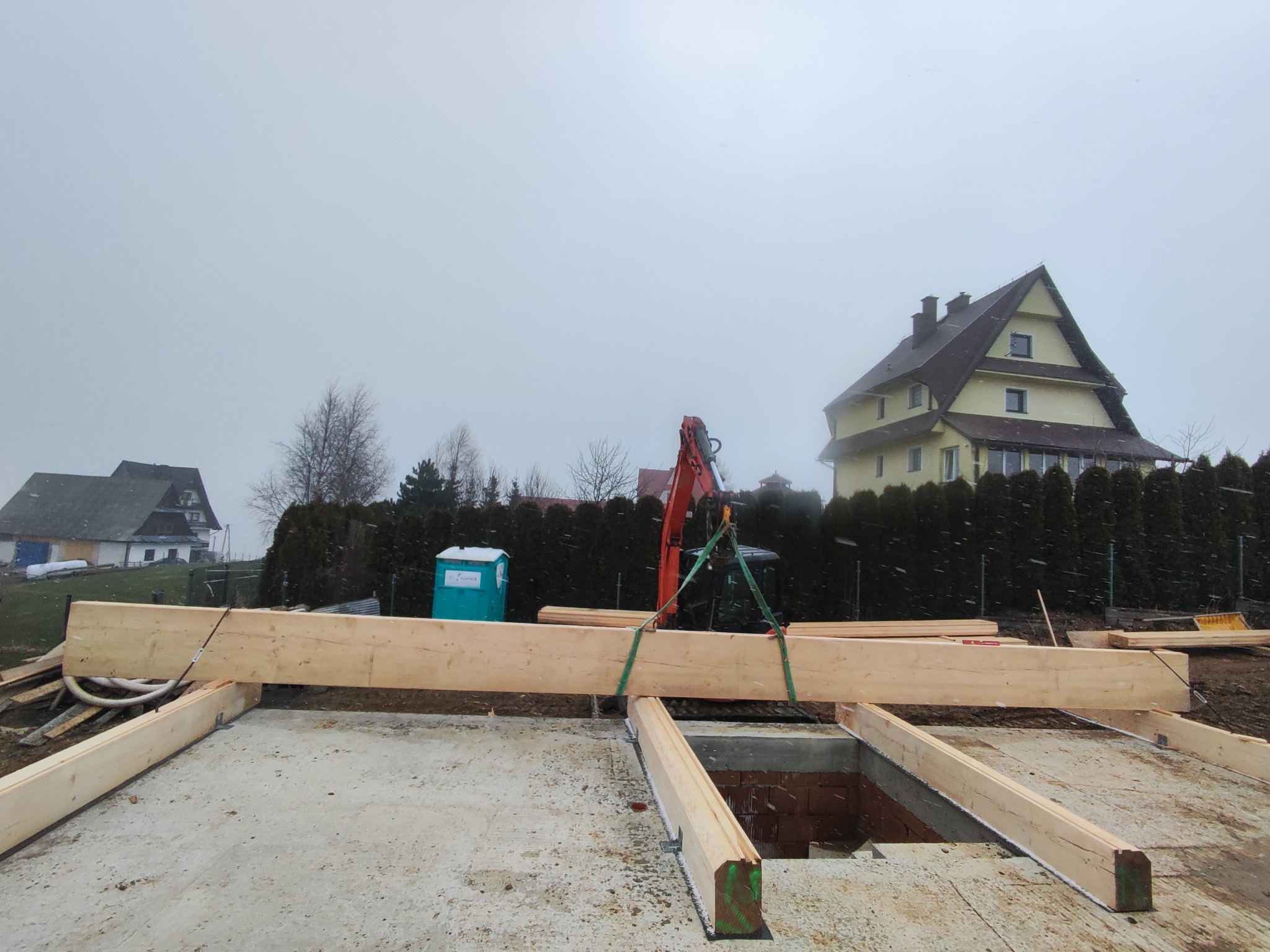 Montaż drewnianych belek stropowych na fundamencie z użyciem dźwigu, widoczne ceglane ściany fundamentowe i budynki w tle, pochmurna pogoda.