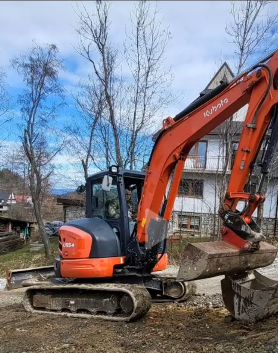 Pomarańczowa mini koparka Kubota U55-4 na nierównym terenie, operator w kabinie, widoczny fragment domu w tle.