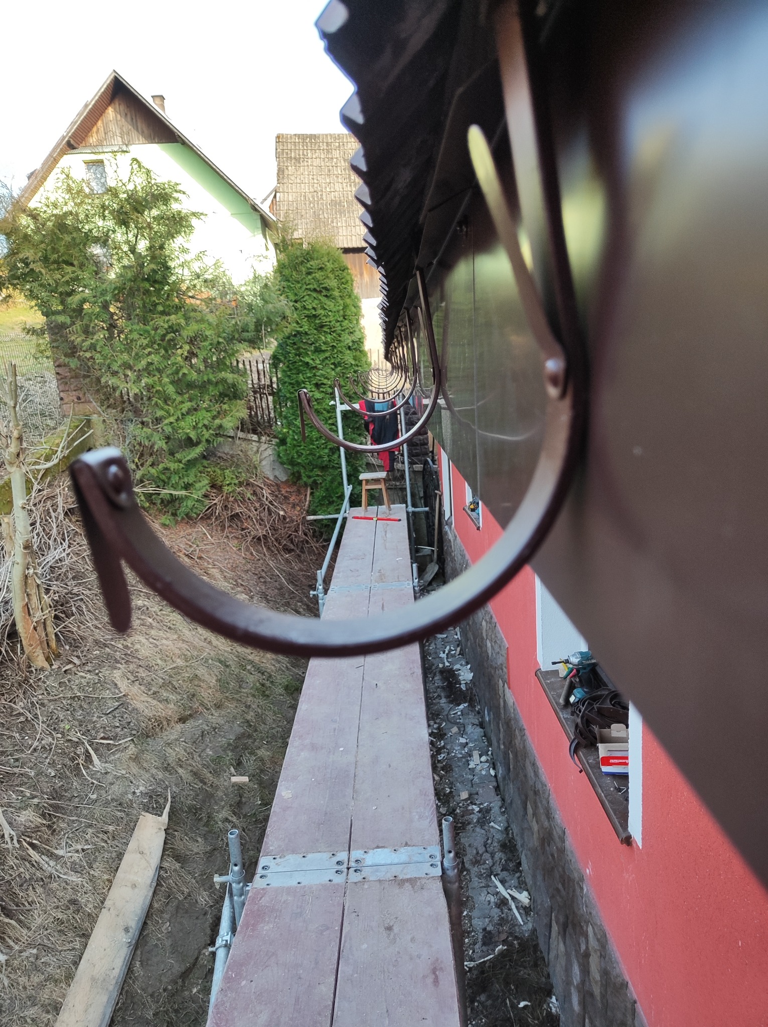 Montaż systemu rynnowego z bliska, widok z rusztowania na brązowe haki rynnowe, narzędzia na parapecie okna, dom w tle.