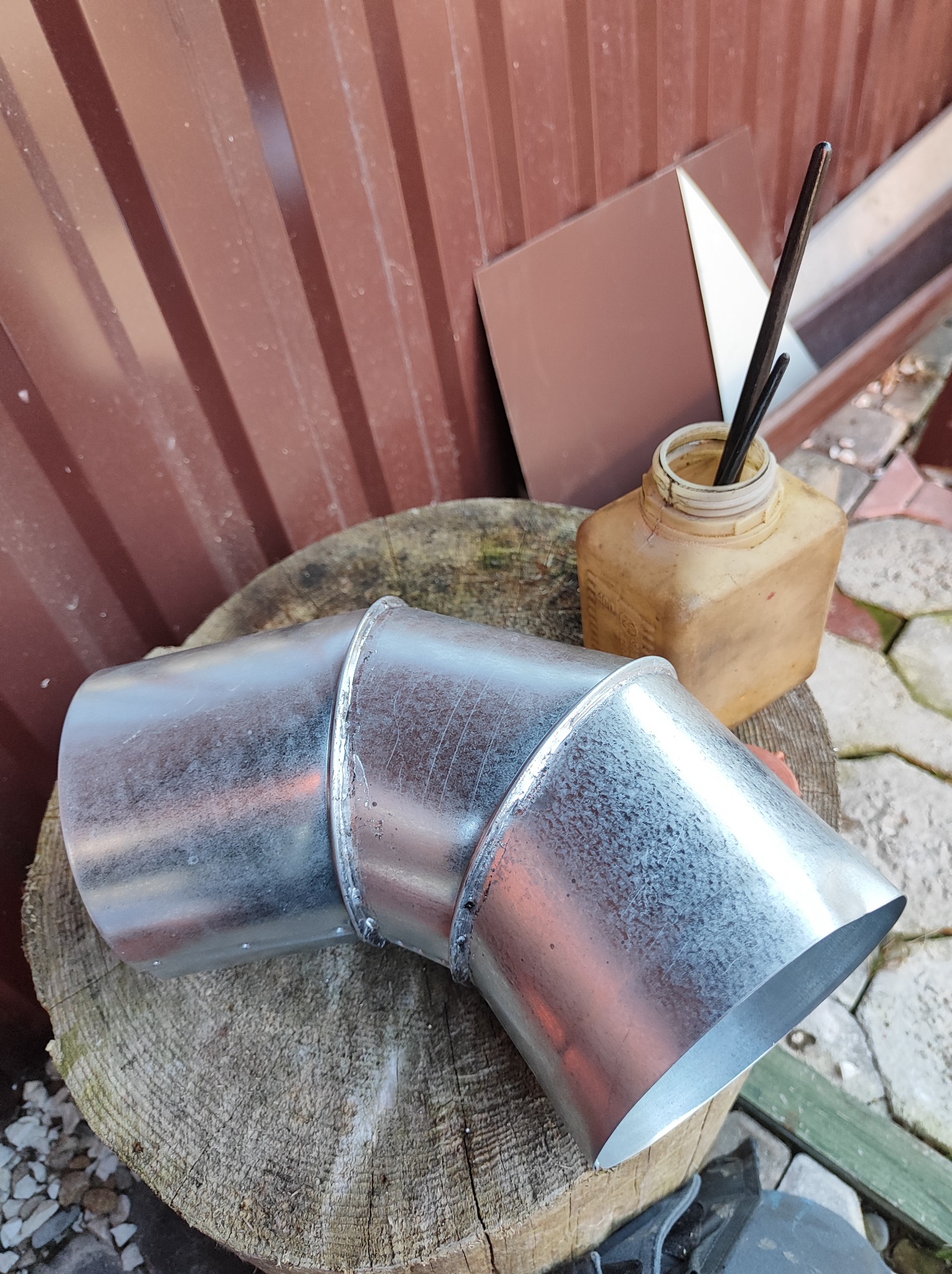 Kolanko rury spustowej z blachy ocynkowanej, leżące na pniu drzewa, obok puszka z pędzlami i próbki blachy w odcieniach brązu, tło stanowi blaszana ściana.