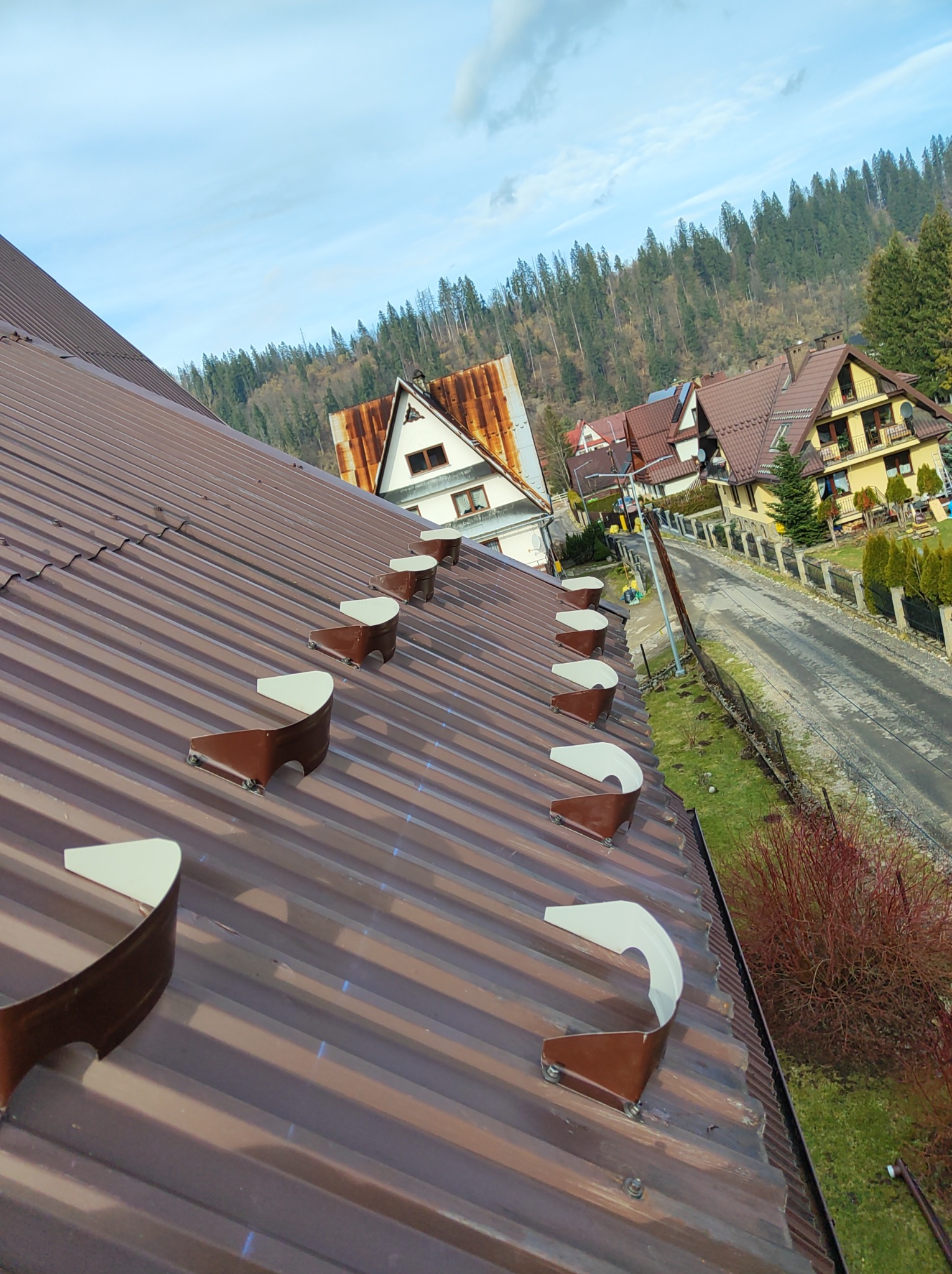 Brązowy blaszany dach z zamontowanymi odśnieżnikami, widok na okoliczne domy i zalesione wzgórza w tle.